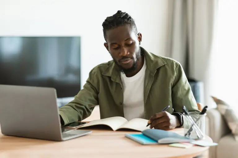 A man working on a laptop from his living room table and writing notes in a notebook