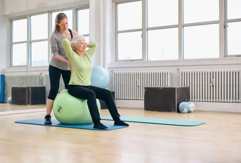 Senior woman sitting on inflatable ball with personal trainer instructing.