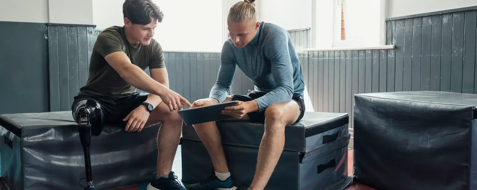 What Should We Start With Today? Young male with prosthetic leg sat talking to personal trainer with digital tablet.