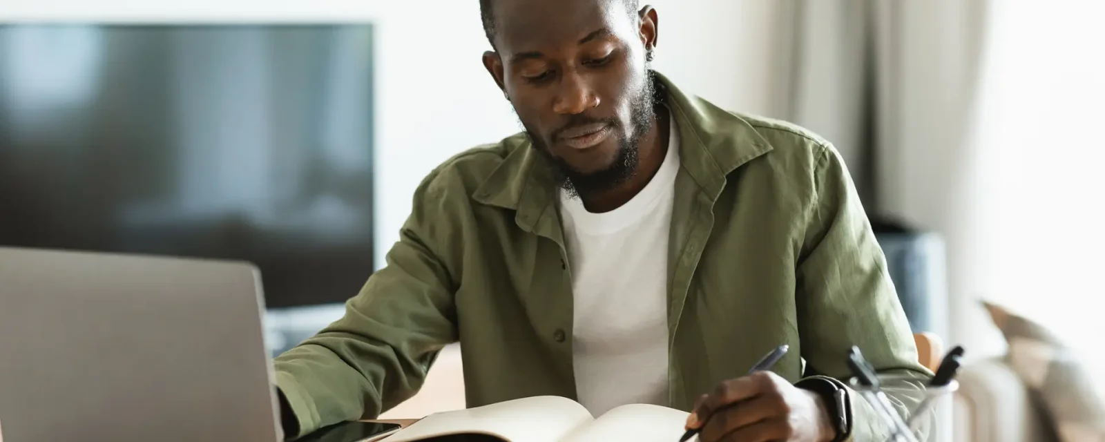A man working on a laptop from his living room table and writing notes in a notebook