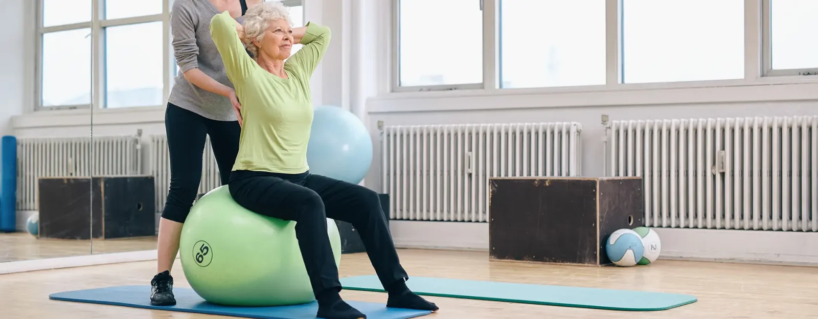 Female trainer helping senior woman exercising in gym Senior woman sitting on inflatable ball with personal trainer instructing.